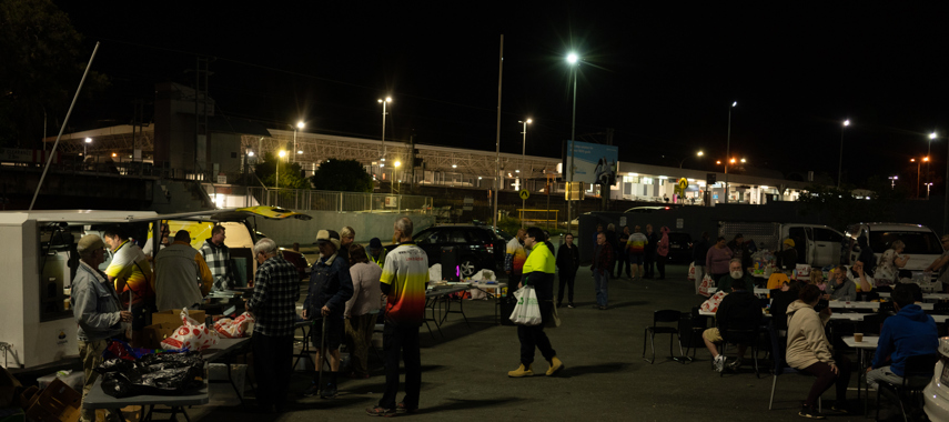 People stand around and help themselves to dinner served by Nightlight each Thursday evening to homeless people beenleigh train station carpart August 2022